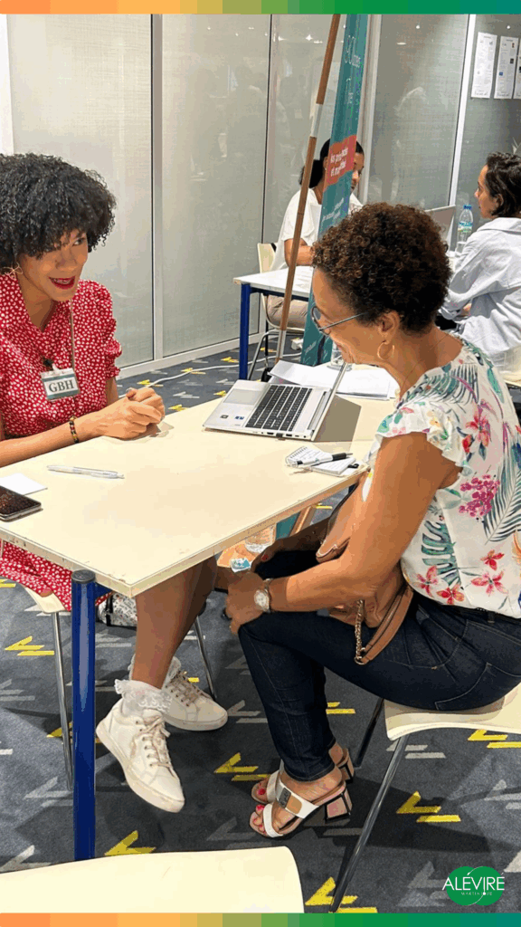 Deux femmes sont assises face à face à une petite table lors d'un JOB Dating Inversé lors du Salon Du Retour. À gauche, une femme portant une robe rouge à pois blancs et des baskets blanches avec des chaussettes engage une discussion attentive. Elle porte un badge avec l’inscription « GBH ». À droite, une autre femme, habillée d’un haut à motifs floraux et d’un jean foncé, écoute attentivement. Elle tient un sac à main camel et est chaussée de sandales à talons ouverts. Sur la table, on aperçoit un ordinateur portable, un bloc-notes, un stylo et un téléphone portable. En arrière-plan, d'autres personnes discutent autour de tables similaires, suggérant une ambiance d’échanges professionnels ou de coaching. En bas à droite de l’image, le logo « ALÉ VIRÉ Martinique » est visible, accompagné d’un cadre coloré dégradé orange-vert en haut et en bas de l’image.
